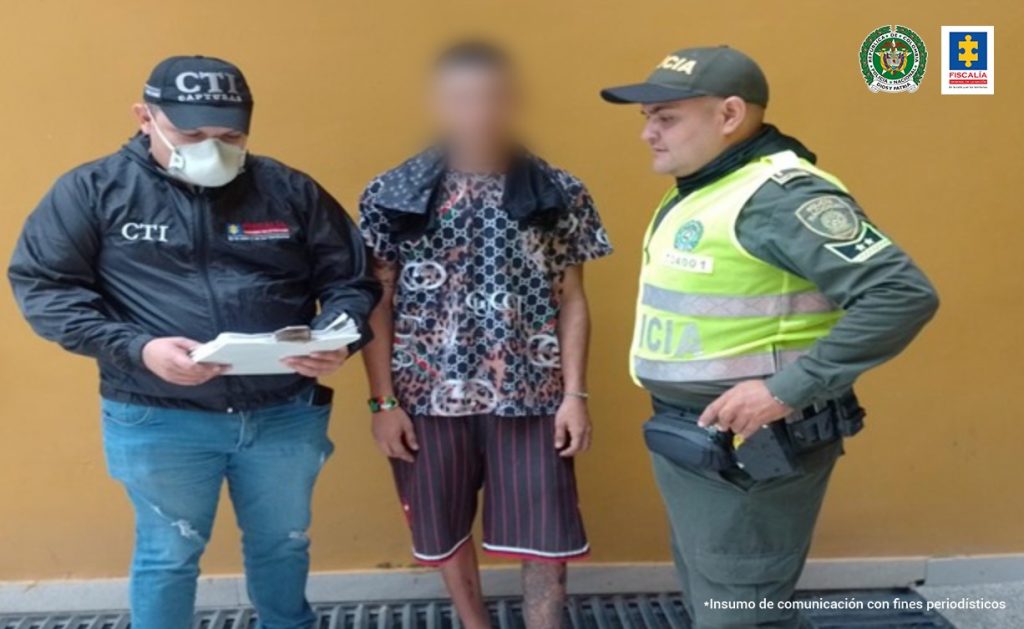 En la imagen se observa a un hombre con el rostro difuminado vestido con camiseta y bermuda oscura custodiado por un uniformado del CTI y un uniformado de la Policía Nacional.