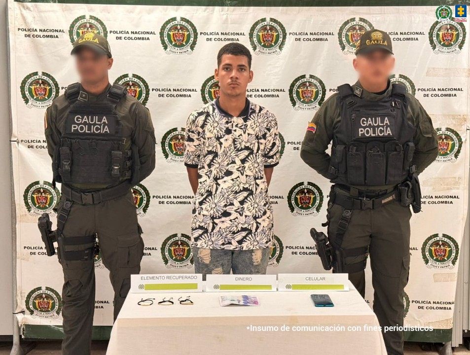 En la fotografía está un hombre joven de pie, moreno, de frente con los brazos atrás esposado, se encuentra vestido con una camiseta de estampados blancos y negros y un jean azul claro. A su lado están dos uniformados de la Policía Nacional y delante una mesa con el dinero y las pulseras de oro incautadas. 