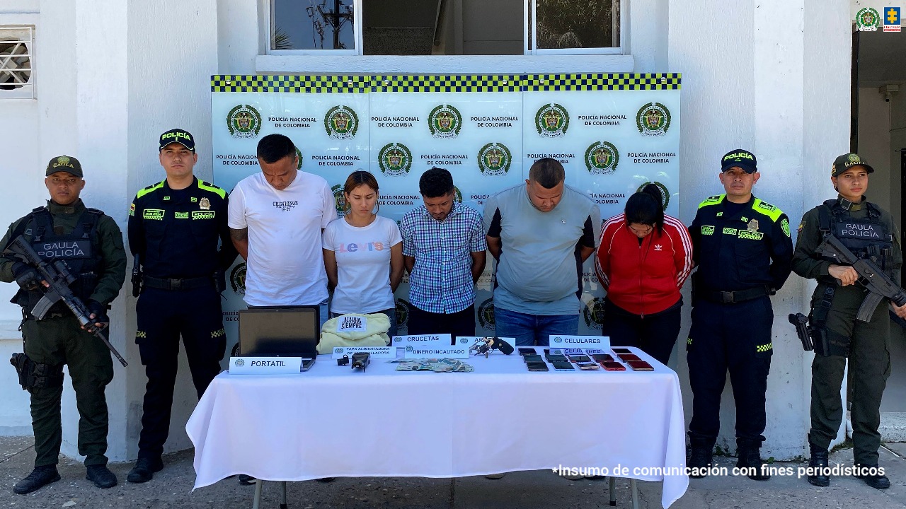 En la fotografía está un hombre joven de pie, moreno, de frente con los brazos atrás esposado, se encuentra vestido con una camiseta de estampados blancos y negros y un jean azul claro. A su lado están dos uniformados de la Policía Nacional y delante una mesa con el dinero y las pulseras de oro incautadas.