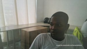 En la foto se ve el medio cuerpo de un hombre moreno, de ojos, nariz, boca y orejas grandes, cabellos cortos, negros, crespos. Está vestido con una camiseta blanca y se encuentra al interior de una oficina asistiendo virtualmente a la audiencia en la que se le procesó. 