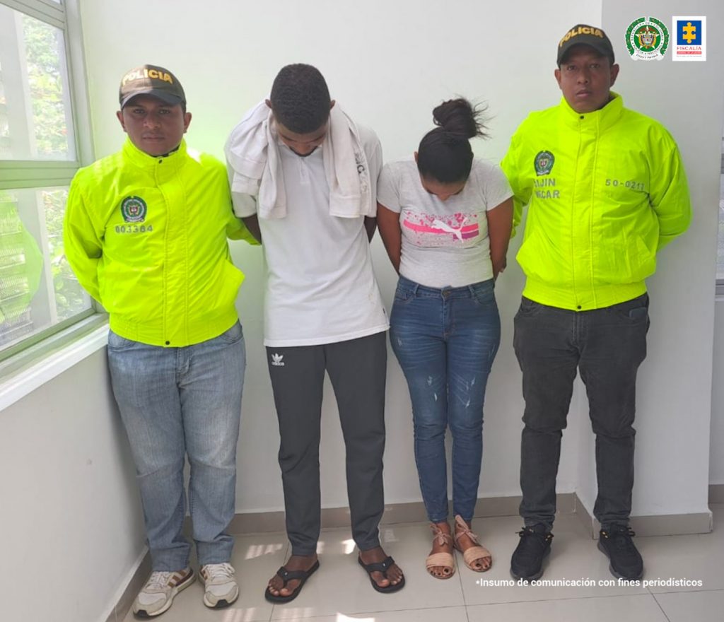 En la imagen se observa a un hombre vestido de camiseta blanca y sudadera gris y a una mujer de camiseta blanca con jean, ambos de frente pero con la cabeza agachada, custodiado por dos agentes de la Policía Nacional.