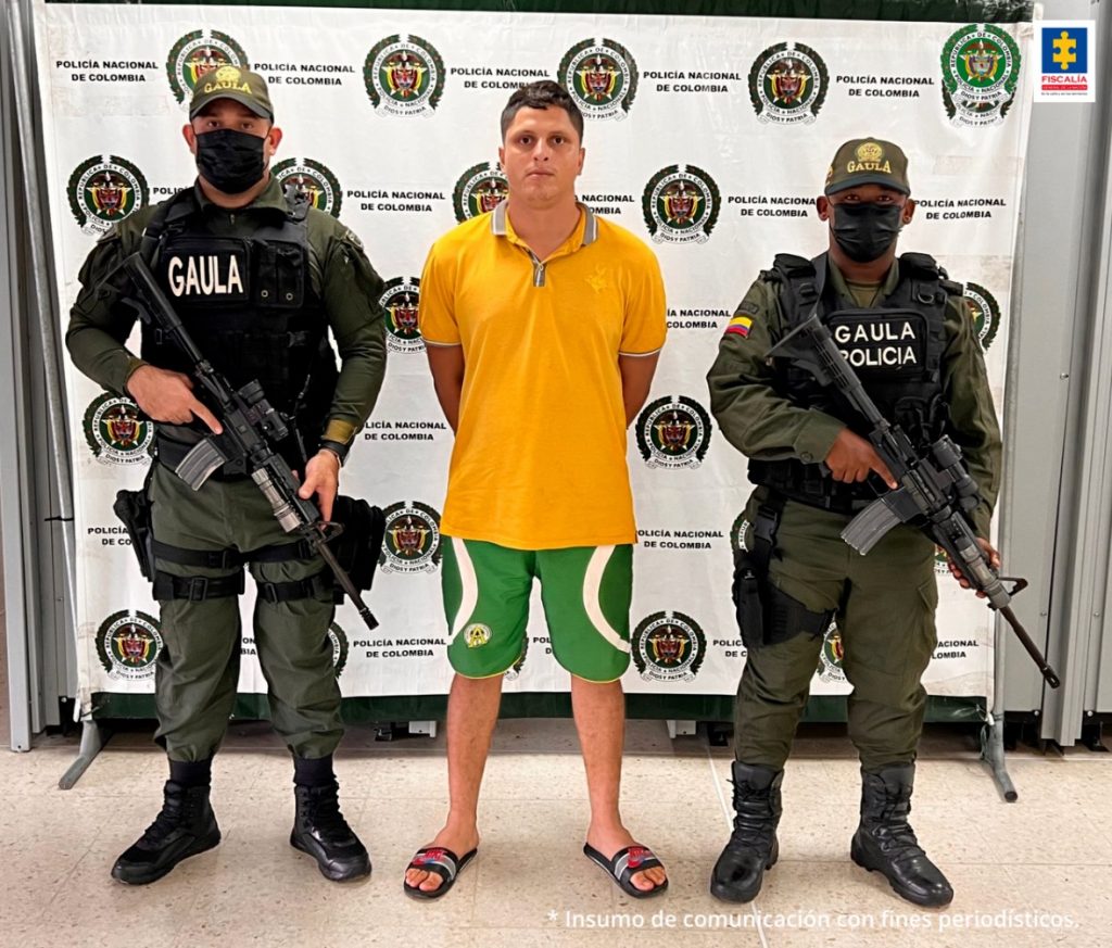 En la imagen se observa a un hombre de tez blanca vestido con camiseta amarilla, bermuda verde y sandalias negras, custodiado por dos agentes de la Policía Nacional delante de un pendón de esa institución.