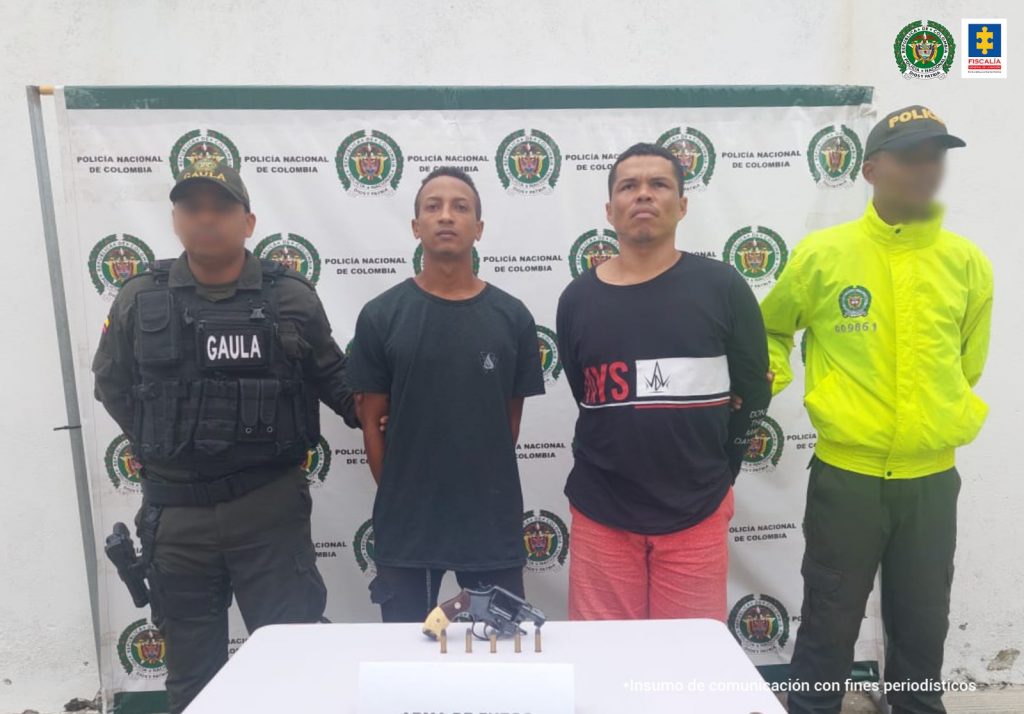 En la fotografía aparecen dos hombres vestidos con camisetas oscuras, custodiados por la Policía Nacional. Detrás de ellos hay un pendón de esa institución y delante un mesa con mantel blanco en la que se encuentra un arma de fuego.