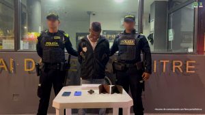En la fotografía esta un hombre de pie, de frente, moreno, con los brazos atrás, cabellos cortos crespos negros, está vestido con una chaqueta negra, camiseta blanca y jean gris. Se encuentra custodiado por dos uniformados de la Policía Nacional.