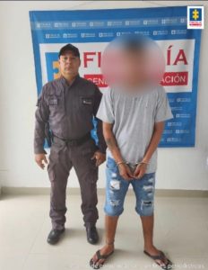 En la fotografía aparece un hombre con el rostro borroso, vestido de camiseta gris, bermuda de jean y sandalias negra con los brazos adelante esposados. A su lado está un funcionario del CTI vestido de uniforme gris y botas negras. Detrás de ellos hay un pendón azul que dice Fiscalía General de la Nación.