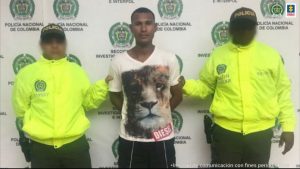 En la fotografía está un hombre joven, moreno de pie, de frente, con los brazos atrás esposados, vestido con una camiseta blanca estampada y un pantalón negro, custodiado por dos uniformados de la Policía Nacional. 