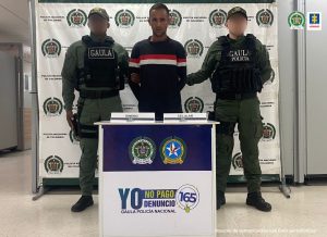 En la fotografía está un hombre de pie, moreno, con los brazos atrás, joven, vestido de buzo azul oscuro con rallas rojas y blancas y pantalón negro, custodiado por dos agentes del Gaula de la Policía Nacional. Detrás de ellos hay un pendón de esa institución.