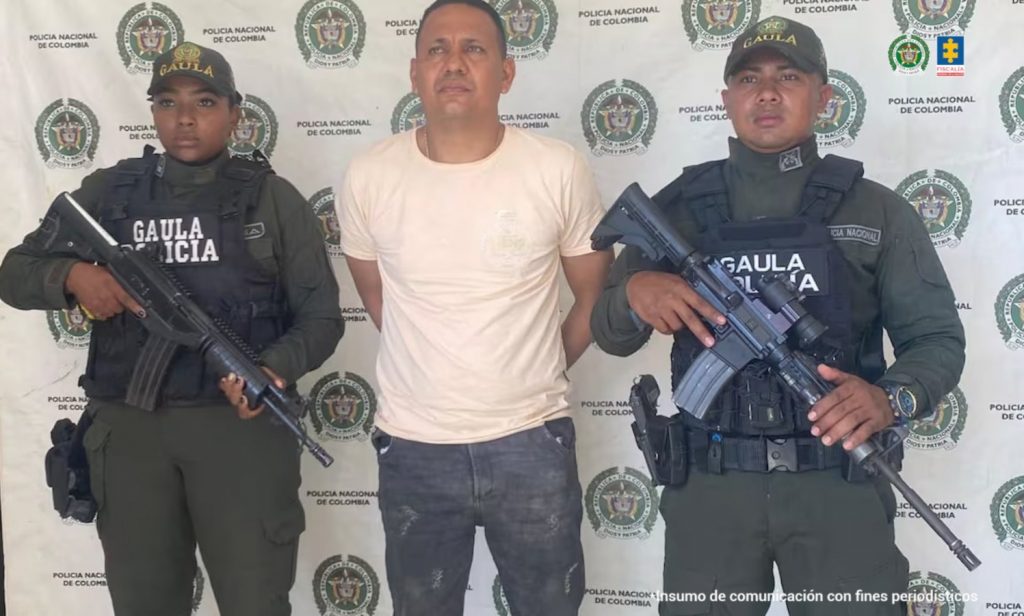 En la fotografía está un hombre alto, trigueño, de pie, con los brazos atrás esposado, cabellos negros, se encuentra vestido con una camiseta beige y un jean gris. Está custodiado por dos uniformados de la Policía Nacional.