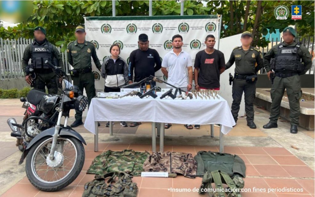 En la fotografía aparecen tres hombres y una mujer de pie, esposados, custodiado por cuatro uniformados de la Policía Nacional. Detrás de ellos hay un pendón de la Policía y delante de ellos una mesa con el material bélico incautado.