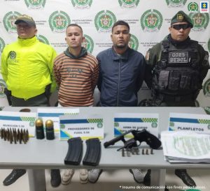 En la fotografía aparecen dos hombres, uno vestido con camiseta de rallas verdes con naranja y el otro de chaqueta azul oscuro custodiados por dos agentes de la Policía Nacional. Delante de ellos hay una mesa donde están un arma de fuego, proveedores y documentos.