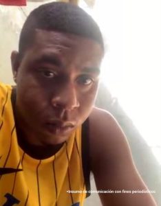 En la fotografía está el rostro de un hombre joven, moreno, de boca, nariz, ojos y orejas grandes, cabellos cortos negros crespos y está vestido con una camiseta amarilla. Se encuentra sentado al interior de una oficina asistiendo virtualmente a la audiencia en la que se le procesó.