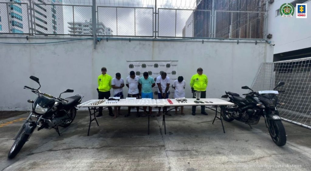 En la fotografía aparecen cinco personas custodiadas por dos agentes de la Policía Nacional delante de un pendón de esa institución. También están unas motocicletas y una mesa con la droga incautada.