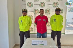En la fotografía está un hombre de pie, con los brazos atrás, de piel trigueña vestido de camiseta roja con jean azul, custodiado por dos agentes de la Policía Nacional.