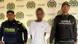 En la fotografía está un hombre de pie, moreno, delgado, con el rostro difuminado, vestido con una camiseta blanca y custodiado por dos uniformados de la Policía Nacional.
