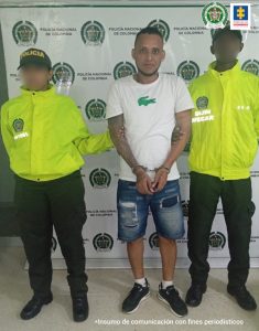 En la fotografía está un hombre de pie, trigueño, con los brazos hacia adelante esposados, vestido de camiseta blanca con verde, bermuda azul de jean, medias blancas y tenis negros. Se encuentra custodiado por dos agentes de la Policía Nacional.