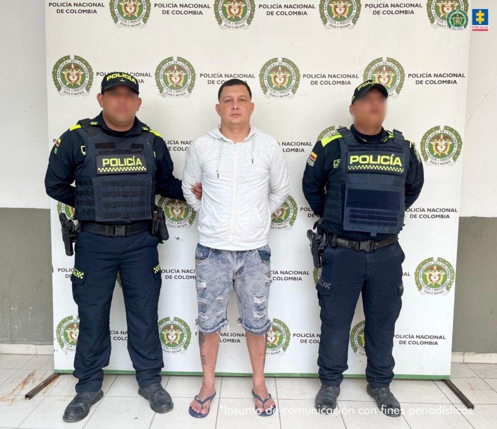 En la imagen está un hombre trigueño de pie, cabellos cortos negros, con los brazos atrás esposados, vestido con un buzo blanco bermuda de jean azul y sandalias oscuras. Se encuentra custodiado por dos uniformados de la Policía Nacional.