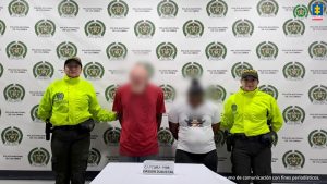 En la fotografía aparecen un hombre de pie, con los brazos atrás, cabeza agachada, rostro difuso, vestido de camiseta roja y jean azul al lado de una mujer de pie, con los brazos atrás, vestida de camiseta blanca y pantalón negro, custodiados por dos agentes de la Policía Nacional.