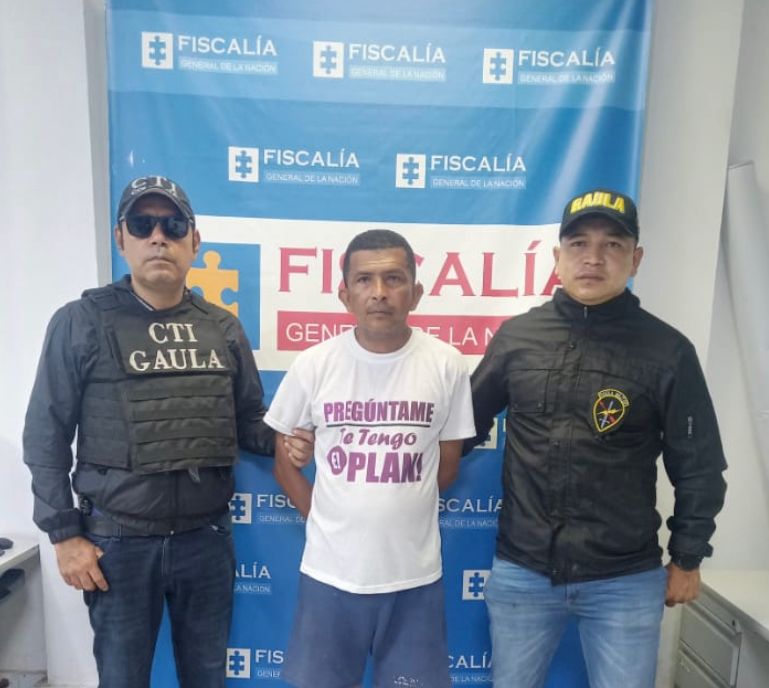 En la imagen se observa a un hombre de camiseta blanca, custodiado por uniformados del CTI de la Fiscalía y del Gaula de la Policía Nacional delante de un pendón institucional de la Fiscalía.