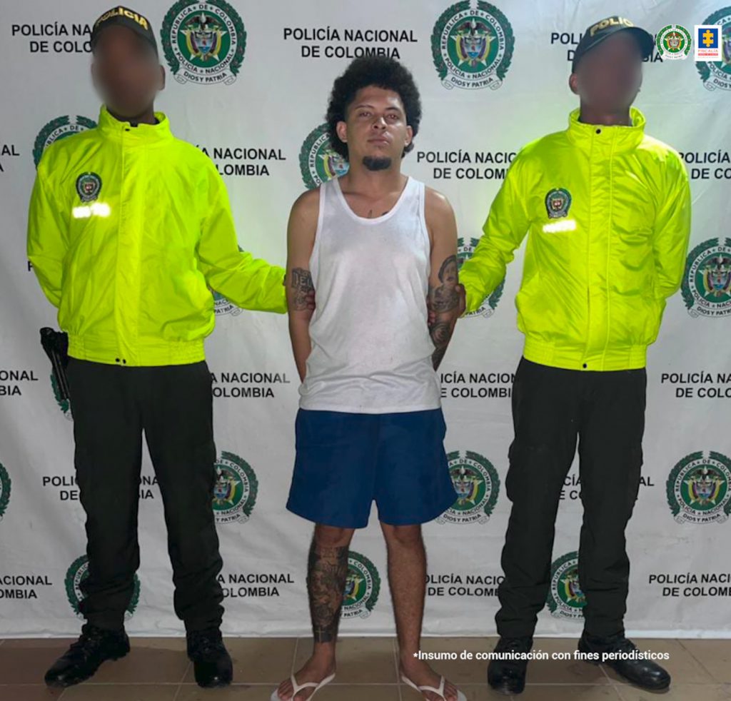 En la fotografía hay un hombre moreno, de pie, de cabellos crespos negro, vestido de camisilla blanca, con bermuda azul y sandalias blancas. Su brazos se encuentran tatuados y su pierna derecha también. Se encuentra custodiado por dos agentes de la Policía Nacional.