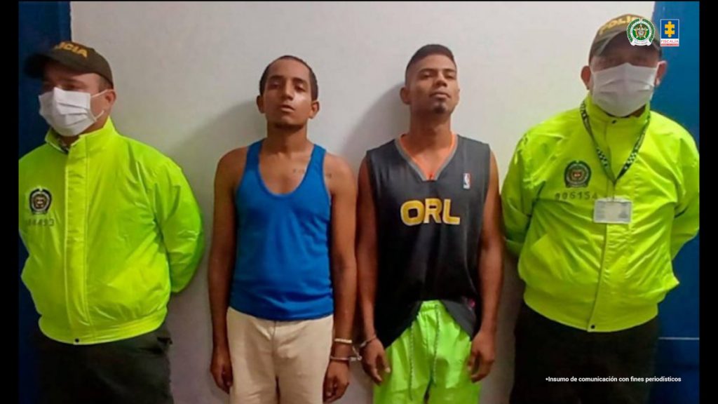 En la fotografía están dos hombres jóvenes de pie, de frente, con los brazos esposados, morenos, delgados, uno está vestido con camiseta azul y bermuda beige y el otro con camiseta negra y bermuda verde. Se encuentran custodiados por dos uniformados de la Policía Nacional. 