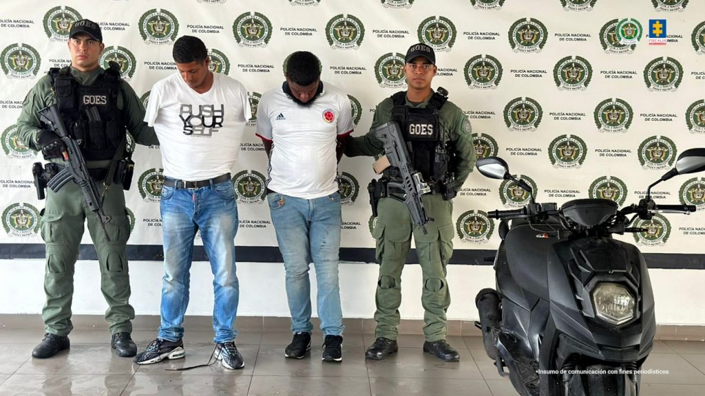 En la fotografía están dos hombres de pie, de frente, morenos, con las cabezas agachadas, de contexturas medianas, ambos vestidos con camisetas blanca y jean azul. Se encuentran custodiados por dos uniformados de la Policía Nacional. 
