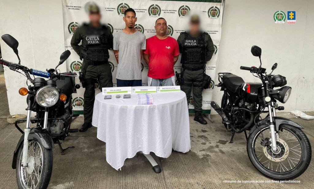En la fotografía están dos hombres de pie, con los brazos atrás esposados, uno es de contextura delgada el otro es de contextura gruesa con abdomen abultado. Se encuentran vestidos con camiseta gris y camiseta roja respectivamente y están custodiados por dos uniformados de la Policía Nacional.