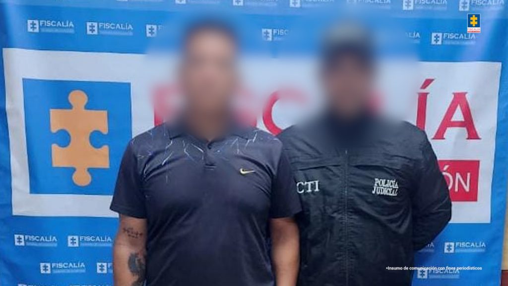 En la fotografía está un hombre de pie, alto, trigueño, cabellos negros, vestido con camiseta oscura, jean y sandalias negras. Se encuentra custodiado por un uniformado del CTI y con el pendón de la Fiscalía detrás de ellos. 