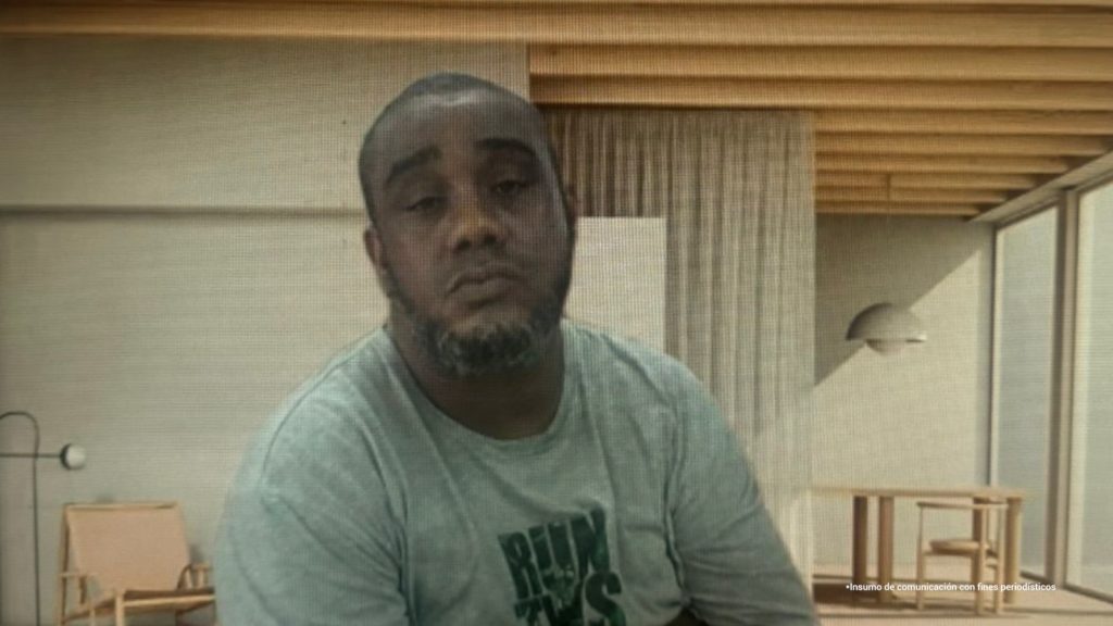 En la fotografía está el medio cuerpo de un hombre moreno, robusto, de cabellos cortos crespos, tiene barba, boca y nariz grande. Se encuentra vestido con una camiseta gris y está sentado en el interior de una sala asistiendo virtualmente a la audiencia en la que se le procesó. 