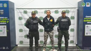 En la imagen está un hombre alto, trigueño, de pie con los brazos atrás, vestido con una chaqueta negra y un pantalón camuflado de color gris con blanco, custodiados por dos antes del Gaula de la Policía Nacional.