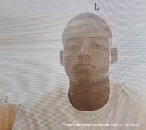 En la fotografía aparece un hombre joven, moreno, delgado, con cabellos cortos, ojos, nariz y boca mediana, vestido con una camiseta blanca. Se encuentra sentado en una oficina asistiendo virtualmente a la audiencia en la que se le procesó.