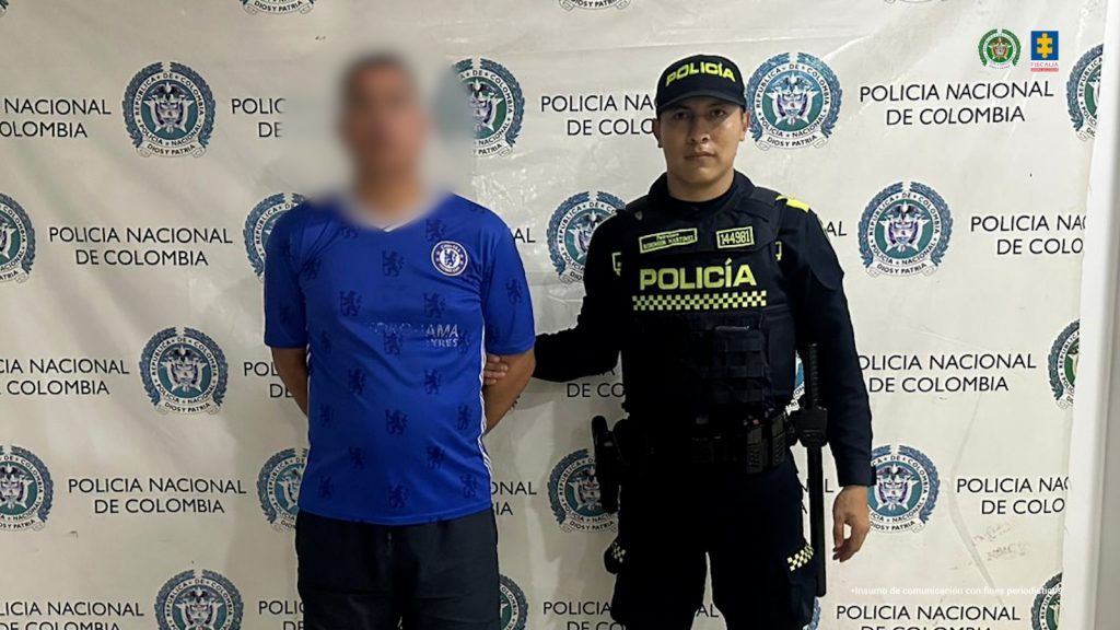 En la foto está un hombre trigueño, de frente, de pie, con los brazos atrás esposados, vestido con una camiseta azul con blanco y un pantalón oscuro. Se encuentra custodiado por un uniformado de la Policía Nacional. 