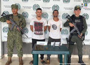 En la imagen están dos hombres de pie, con los brazos atrás, trigueños, vestidos ambos de camiseta blanca y pantalones oscuros, custodiado por uniformados de la Policía y la Infantería de Marina. Delante de ellos hay una mesa verde donde están las municiones incautadas.