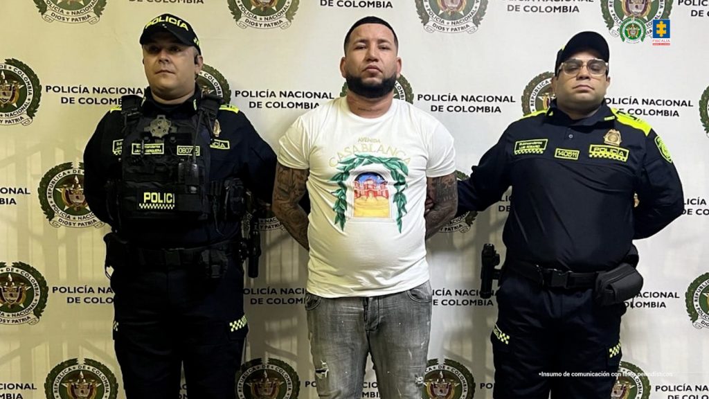 En la imagen está un hombre moreno, de pie, de frente, de contextura mediana, con los brazos atrás esposados, cabellos y barba negra, está vestido con una camiseta blanca y un jean gris. Se encuentra custodiado por dos uniformados de la Policía Nacional. 