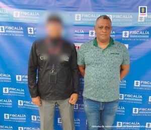 En la imagen está un hombre vestido de pie, con los brazos atrás, vestido con camiseta verde y jean azul, custodiado por un funcionario del CTI. Detrás de ellos se encuentra un pendón azul de la Fiscalía General de la Nación.