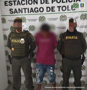 En la fotografía hay un hombre que se encuentra de pie, con el rostro difuso y los brazos atrás. Está vestido con camiseta rosada y bermuda de jean azul, custodiado por dos agentes de la Policía Nacional.  Detrás de ellos hay un pendón de esa institución. 