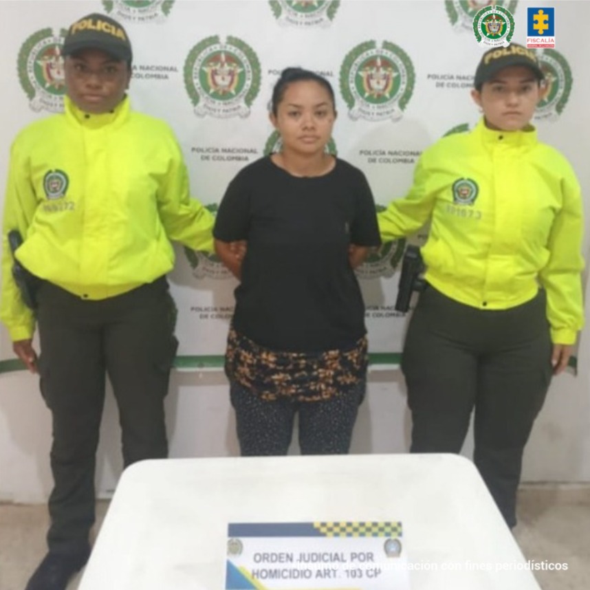 n la imagen, está una mujer de pie, morena, con los brazos atrás, de contextura mediana, estatura baja, cabellos negros recogidos, vestida con camiseta negra y pantalón negro con puntos blanco, custodiada por dos uniformadas de la Policía Nacional.