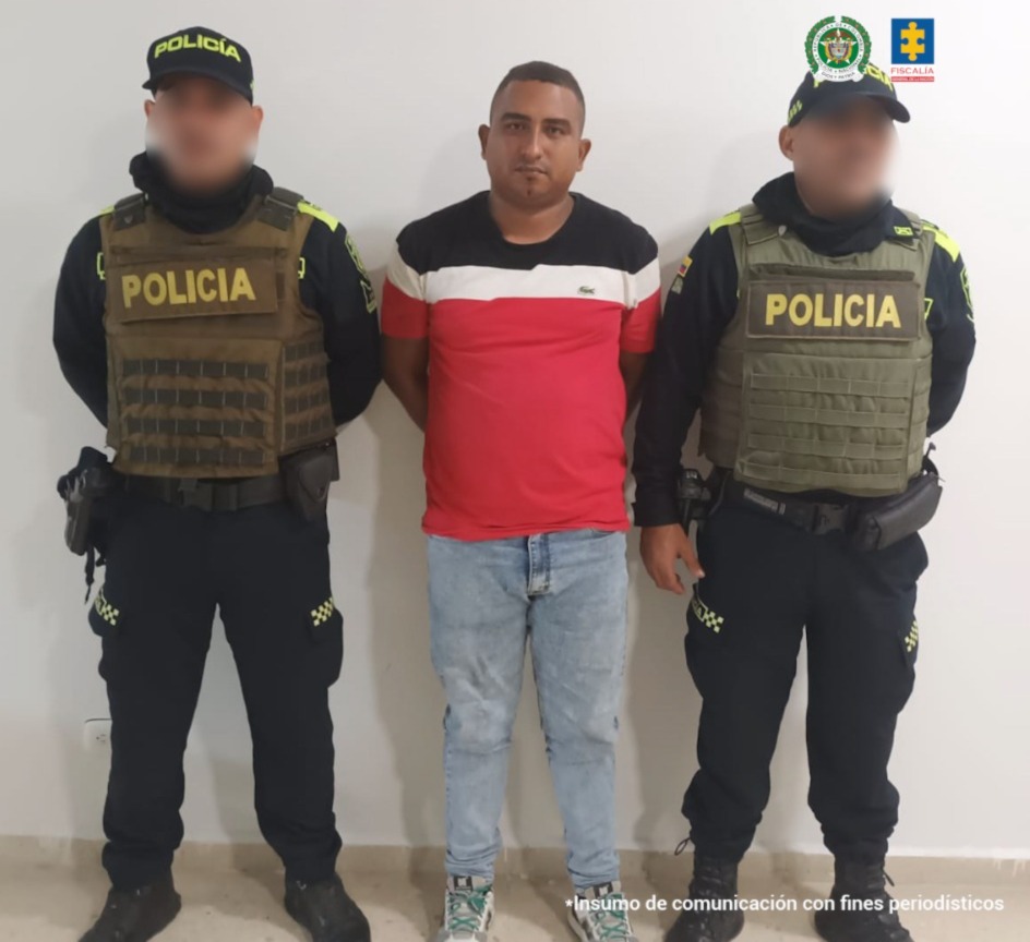 En la fotografía, aparece un hombre de pie, moreno, con los brazos atrás, de contextura y estatura mediana, vestido con una camiseta roja con rallas blanca y azul, tenis blanco con negro y cordones verdes, custodiado por dos uniformados de la Policía Nacional.
