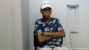 En la imagen se observa a un hombre sentando con gorra blanca de camiseta azul oscura con blanco, tiene los brazos cruzados. A su lado se aprecia también una muleta.