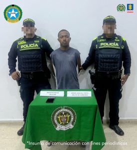  En la fotografía está un hombre de frente, de pie, de baja estatura, con los brazos atrás esposados, cabellos cortos crespos, labios y nariz gruesas y se encuentra vestido con una camiseta azu. A su lado se encuentran dos uniformados de la Policía Nacional. 