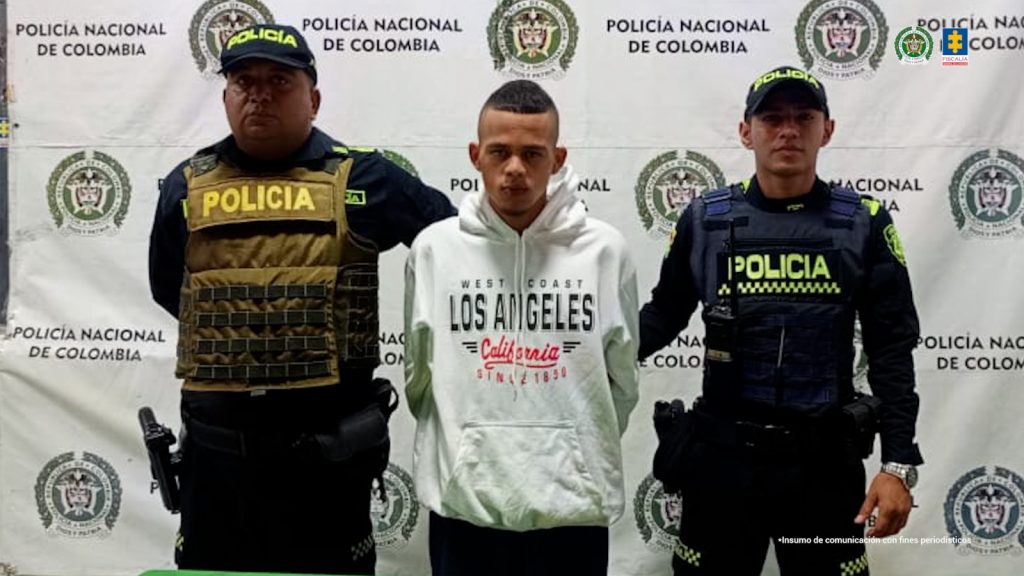En la fotografía está un hombre joven, delgado de pie, de frente con los brazos atrás esposados, vestido con un buzo blanco y un pantalón negro. Se encuentra custodiado por dos uniformados de la Policía Nacional. 