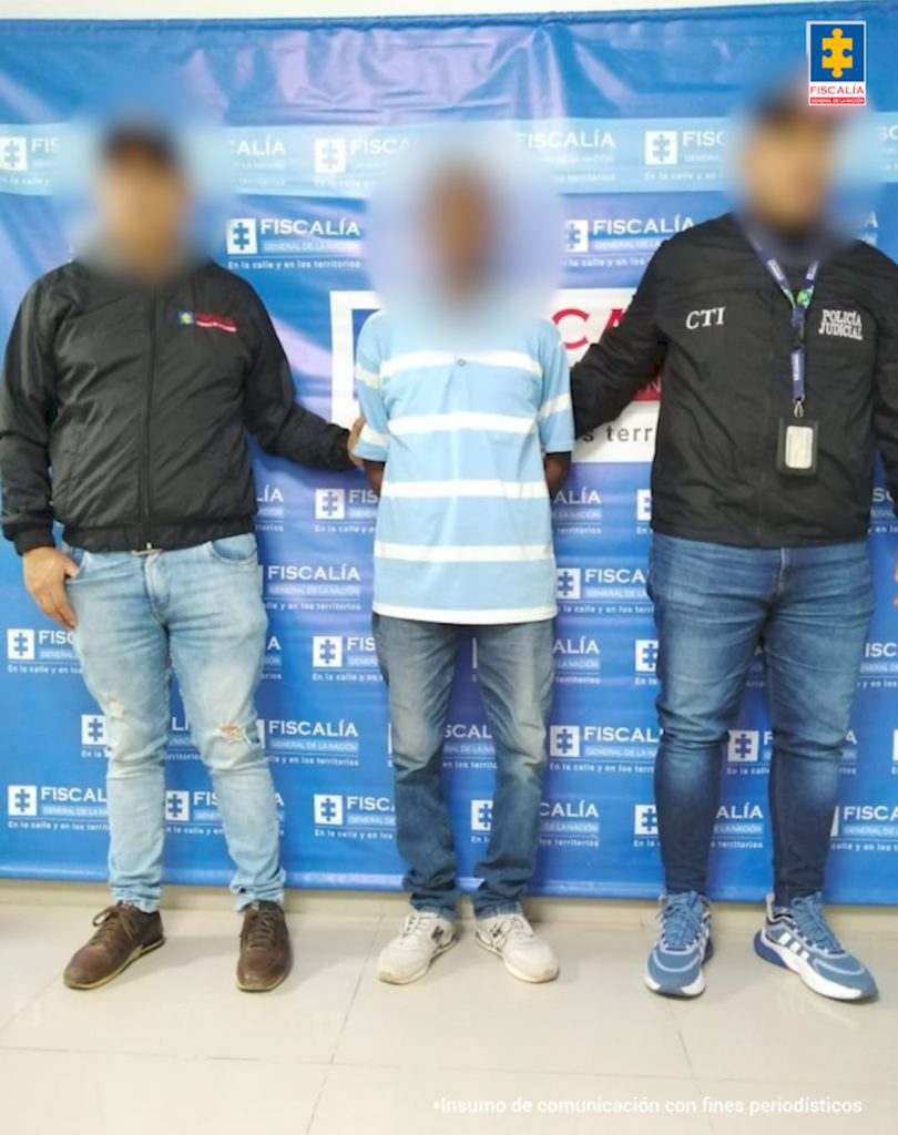 En la fotografía está un hombre de pie, con el rostro difuso, los brazos atrás, vestido de camiseta azul con rallas blancas, jean azul y tenis blanco, custodiado por dos funcionarios del CTI. Detrás de ellos hay un pendón azul de la Fiscalía General de la Nación.