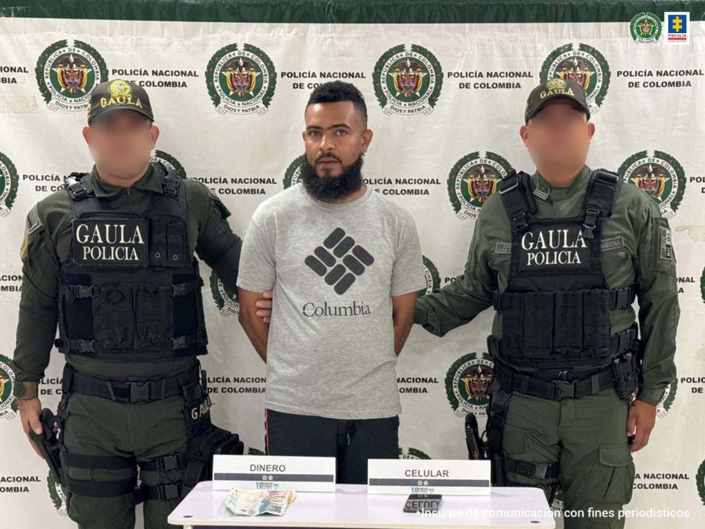 En la fotografía aparece un hombre moreno, de pie, con los brazos atrás, de cejas unidas, barba negra, vestido con una camiseta gris y un pantalón negro, custodiados por dos uniformados de la Policía Nacional. Delante de ellos esté el dinero que se le incautó.