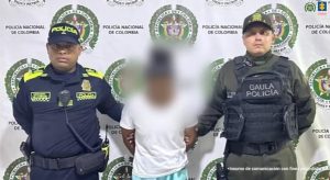 En la imagen está el medio cuerpo de una mujer morena, de pie, de frente, con los brazos atrás esposados, tiene el rostro difuminado y está vestida con una camiseta blanca y un pantalón azul claro. A su lado se encuentran dos uniformados de la Policía Nacional.