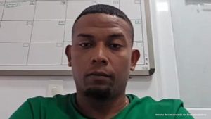 En la fotografía está el rostro de un hombre joven, moreno, cabellos negros cortos negros, vestido con una camiseta verde, se encuentra sentado asistiendo virtualmente a la audiencia en la que se le procesó.