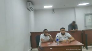 En la imagen están dos hombres morenos, con barbas, cabellos negros crespos, ambos se encuentran sentados en un escritorio de madera con micrófono, vestidos con camisetas blanca en la sala de audiencia donde se les procesó