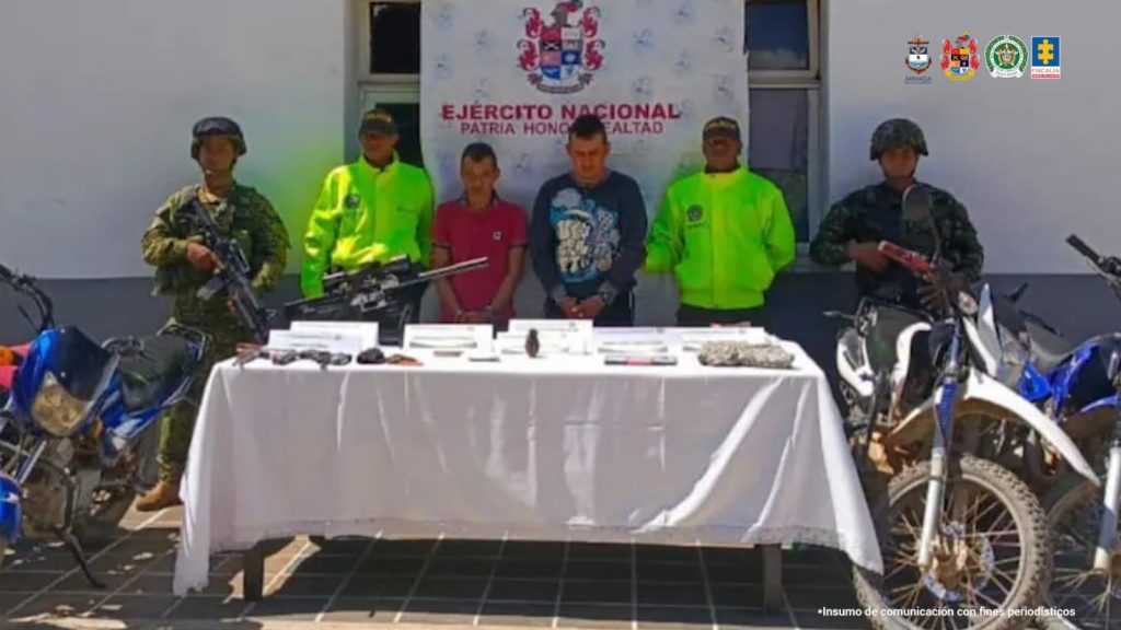 En la fotografía están dos hombres de pie con los brazos adelante esposados, custodiados por uniformados de la Policía, Ejército e Infantería. Delante de ellos hay una mesa con el material bélico incautado.