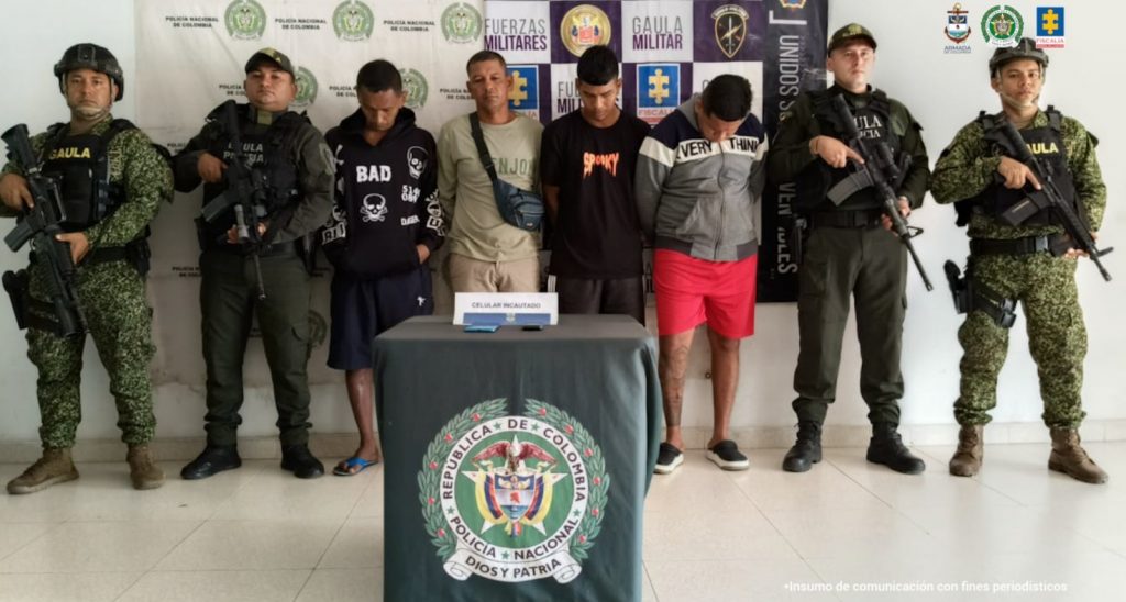 En la imagen están cuatro hombres de pie, con las cabezas agachadas, los brazos atrás, custodiados por uniformados del Gaula de la Policía y la Armada Nacional.