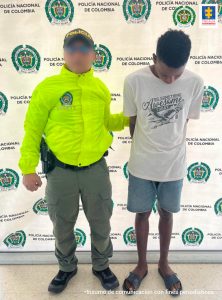 En la fotografía se encuentra un hombre de pie de contextura delgada, trigueño, vestido de camiseta blanca, bermuda gris y sandalias grises, escoltado por un agente de la Policía Nacional.