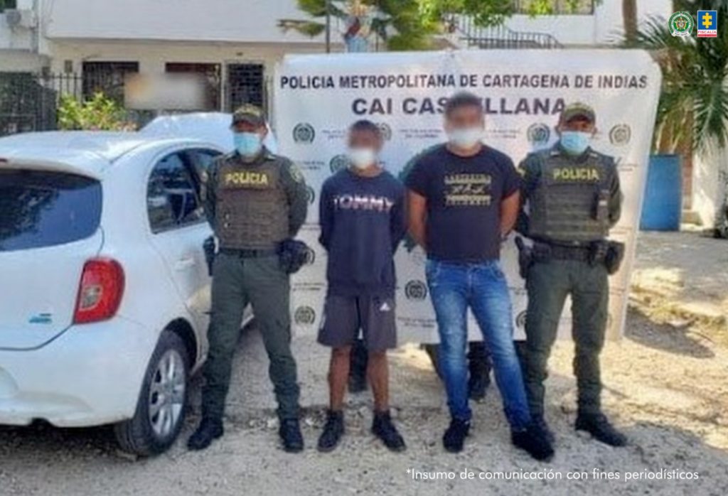 En la fotografía están dos hombres de pie con los brazos atrás esposados, con tapabocas, uno es de baja estatura y está vestido con un buzo negro y una pantalonenta gris y el otro es alto vestido con camiseta negra y jean azul. Se encuentran custodiados por dos uniformados de la Policía Nacional.