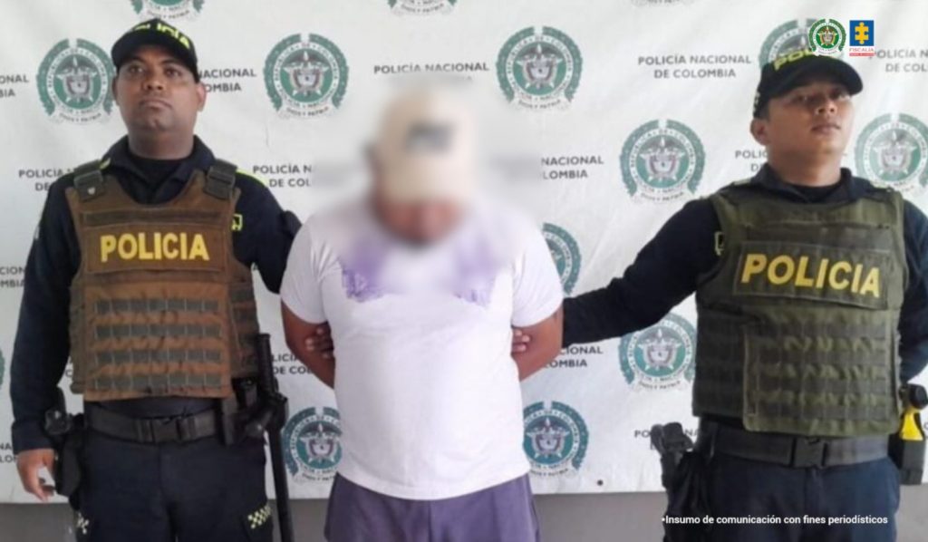 En la fotografía está un hombre trigueño de pie, con una gorra, la cabeza agachada, rostro difuso, vestido con camiseta blanca y pantalón morado. Se encuentra custodiado por dos uniformados de la Policía Nacional.